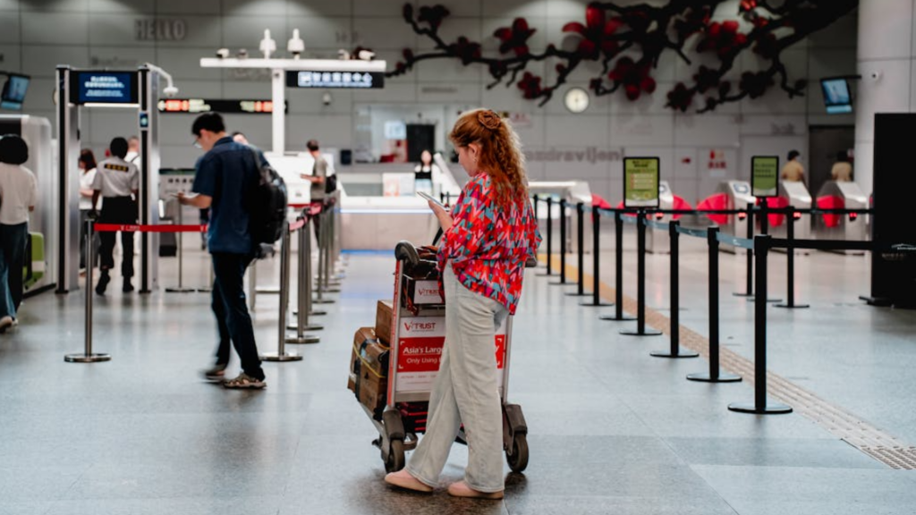 A woman looks at her phone while standing with a luggage cart in a terminal.