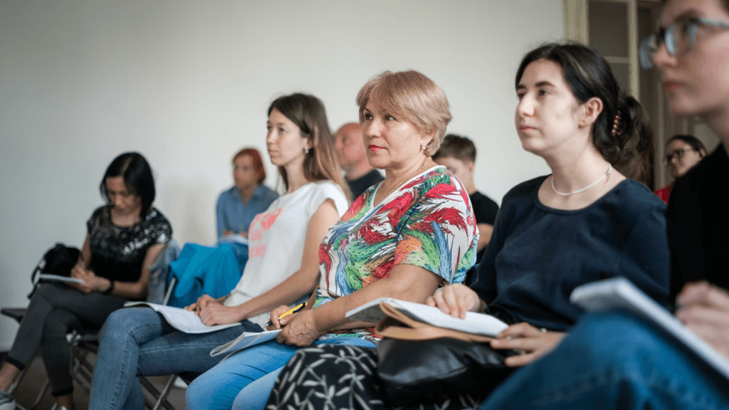 A group of people sit in chairs with notebooks while listening to a meeting.