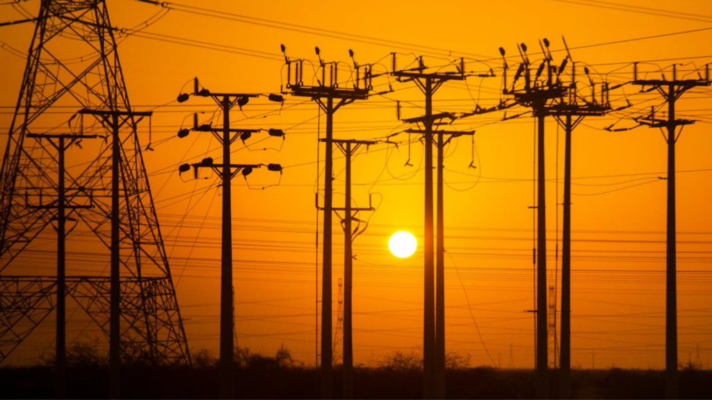 The dark silhouettes of tall electrical power lines and poles stand against a bright orange sunset sky.