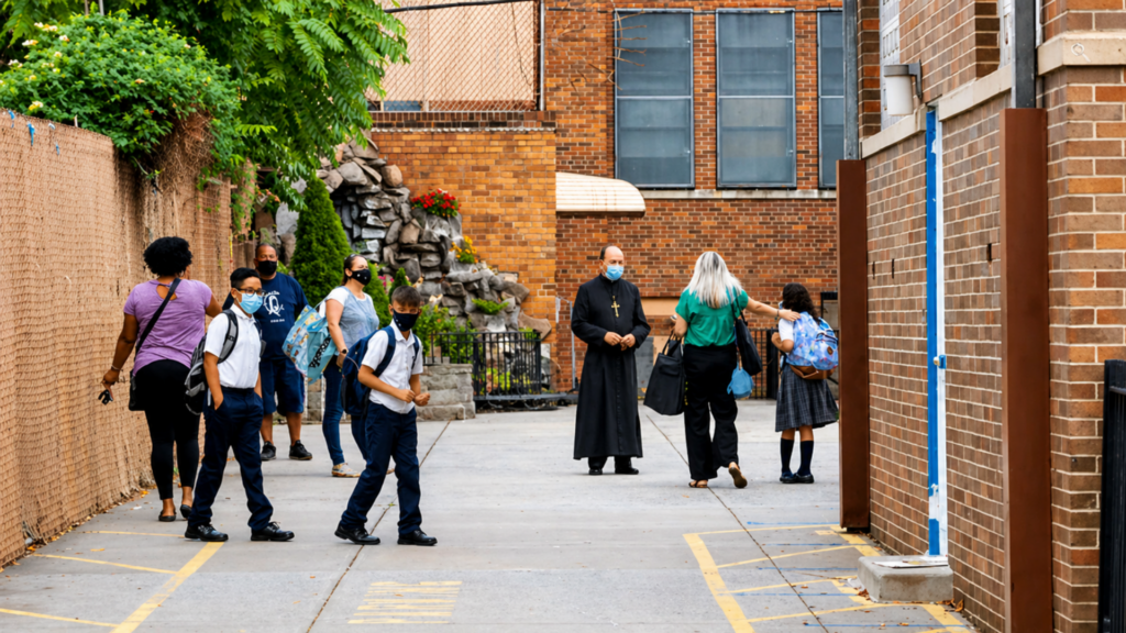 Students and adults wearing masks outside school building with priest nearby.