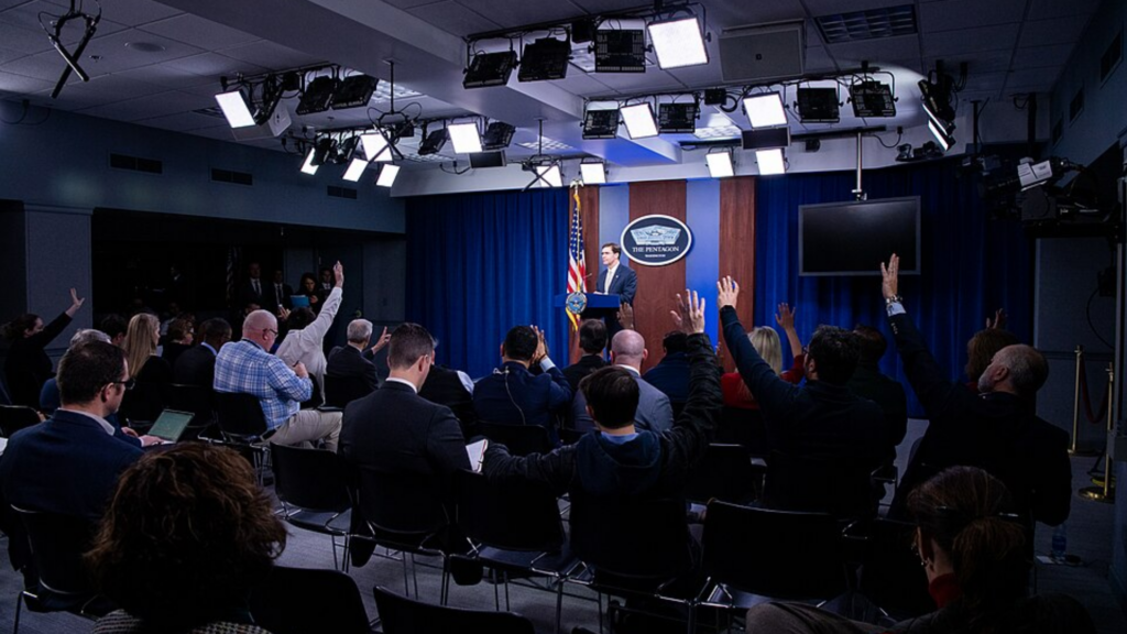 Journalists raise hands during a Pentagon press briefing as a spokesperson speaks at the podium.