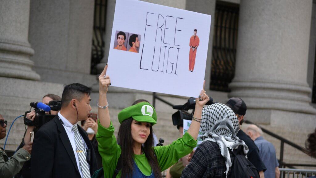 Protester in green Luigi hat holding “Free Luigi” sign outside courthouse with crowd.