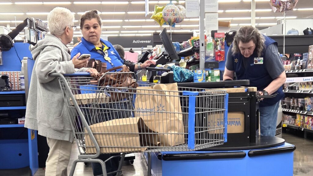 Shoppers and Walmart employees at a checkout lane with a cart full of bagged groceries.
