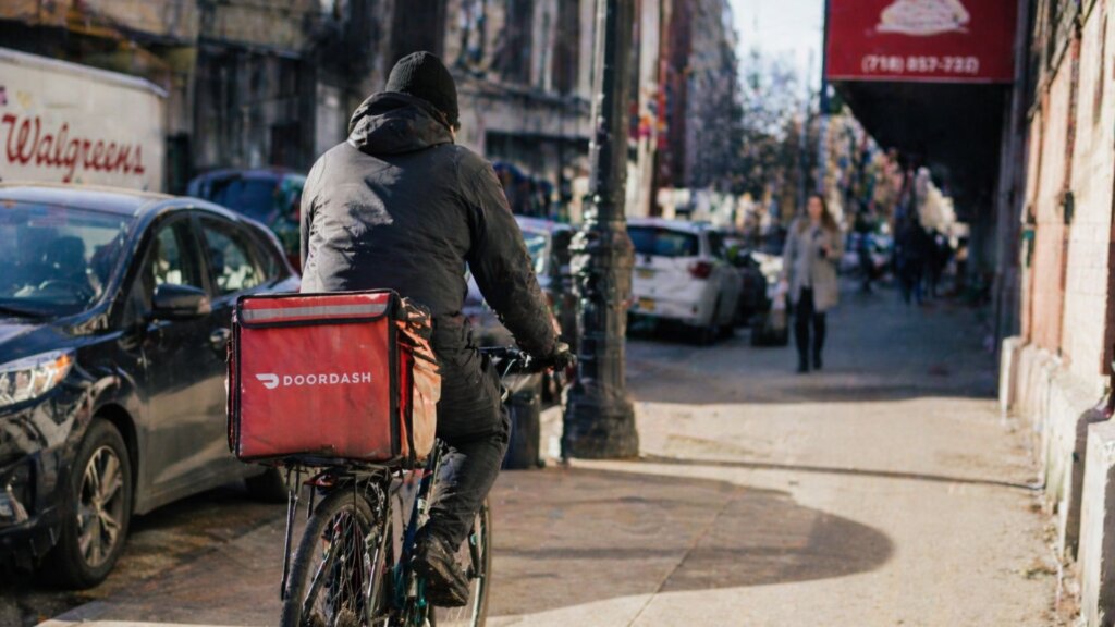A gig economy delivery worker riding a bicycle through a busy city street during the day.