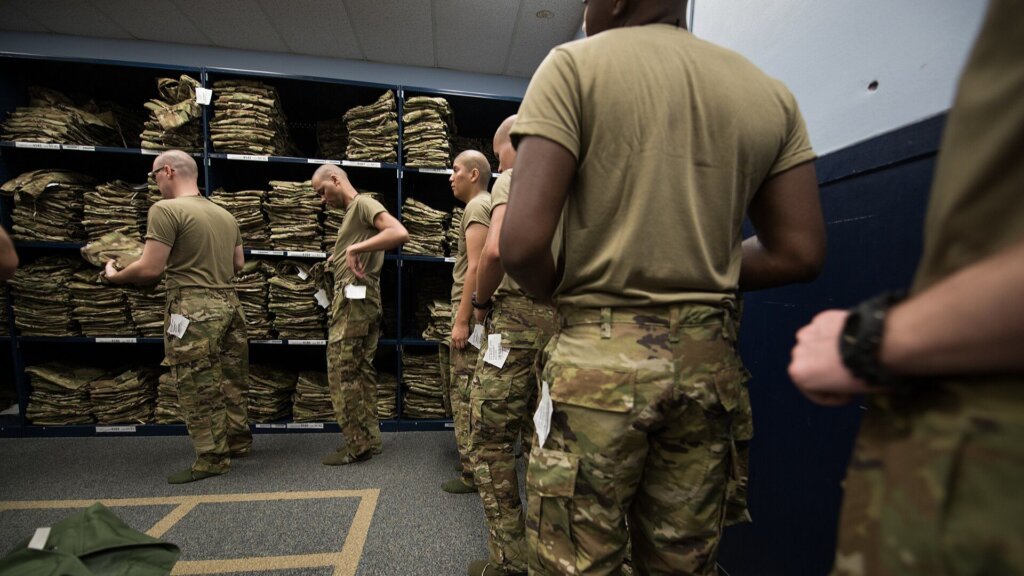 U.S. soldiers in uniform collecting gear from shelves inside a military supply room.