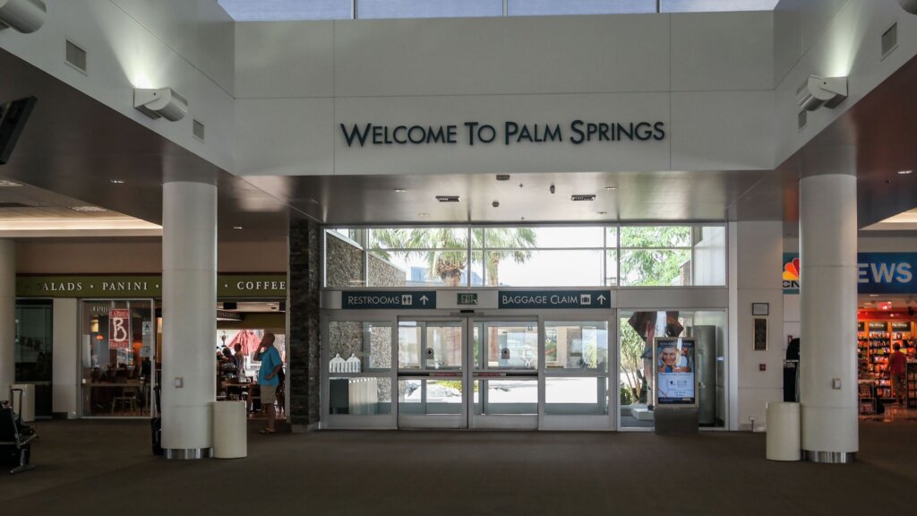 Airport entrance hall with a large sign reading "Welcome To Palm Springs" above glass doors leading outside, with shops and seating areas on either side.