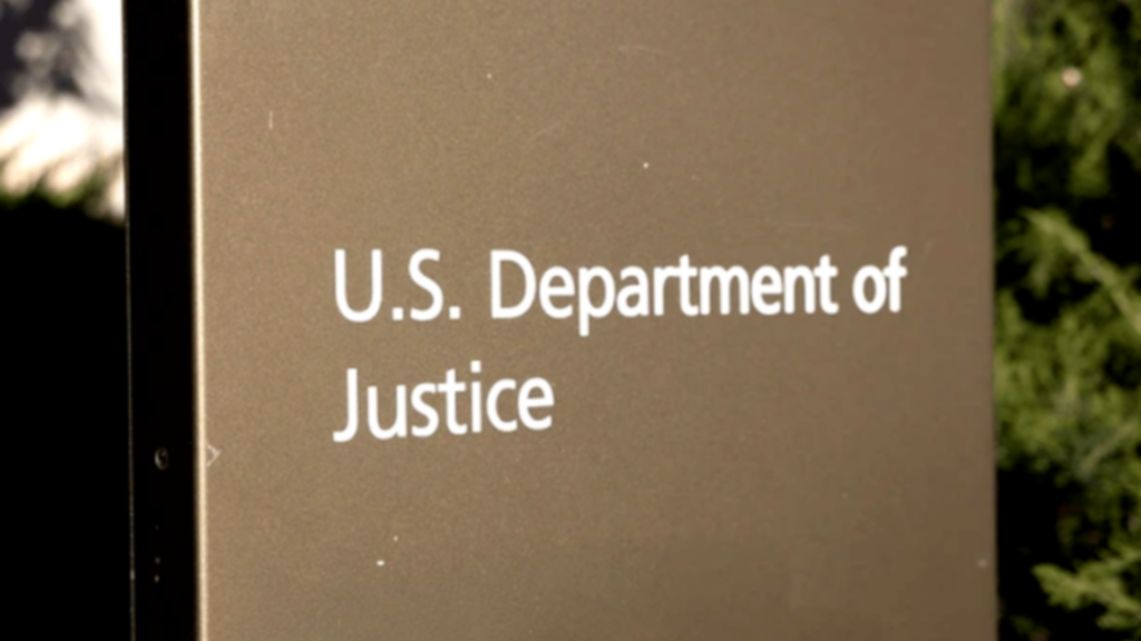 A close-up, slightly angled shot of a gold-toned sign that reads "U.S. Department of Justice".
