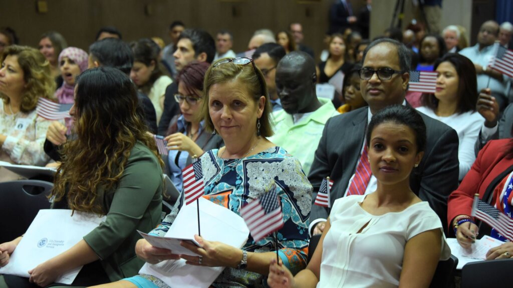 New citizens sit holding U.S. flags during a naturalization ceremony in a crowded room.