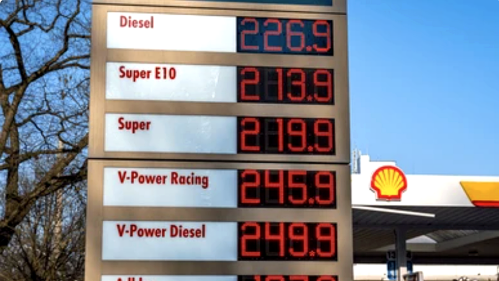 A close-up of a digital fuel price board at a Shell station in Europe; prices are listed in Euros per liter, with Diesel at €2.269, Super E10 at €2.139, and V-Power Diesel at €2.499 under a clear blue sky.