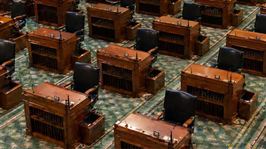 Rows of wooden desks and black chairs arranged across a formal legislative chamber with patterned green carpet.
