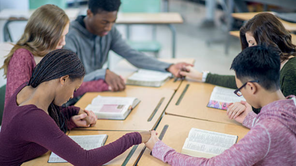 A diverse group of five high school students sitting around a classroom desk, bowed in prayer while holding hands over open books in a quiet, respectful setting.