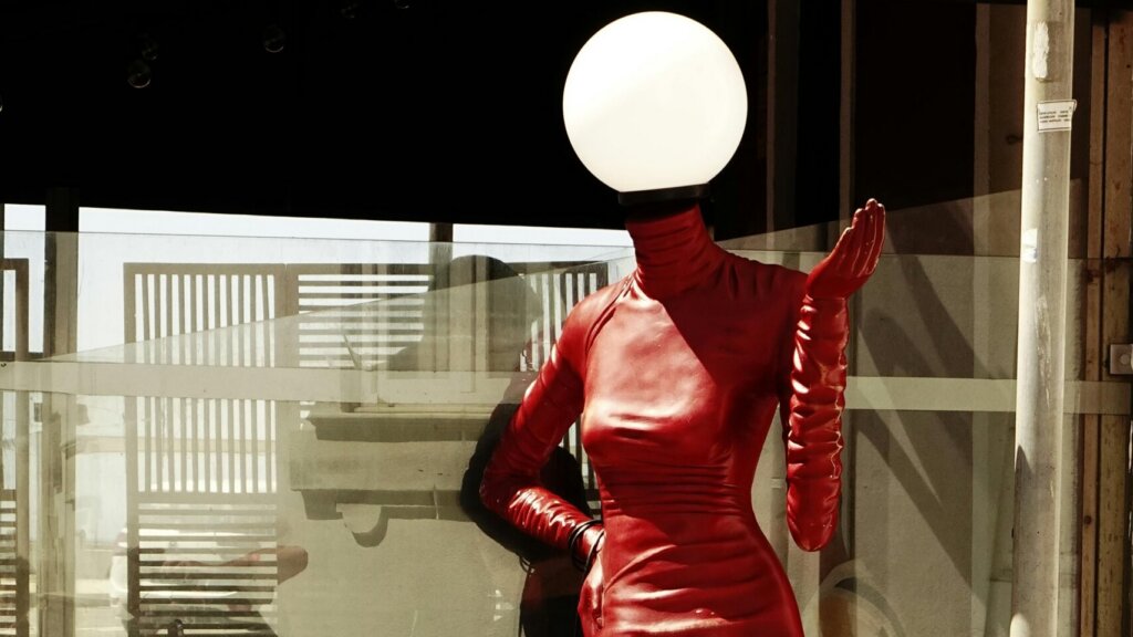 Model in a shiny red latex dress posing in a glass-walled interior setting.