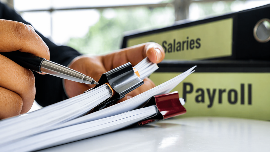 Person holding pen and flipping through documents beside binders labeled Salaries and Payroll.