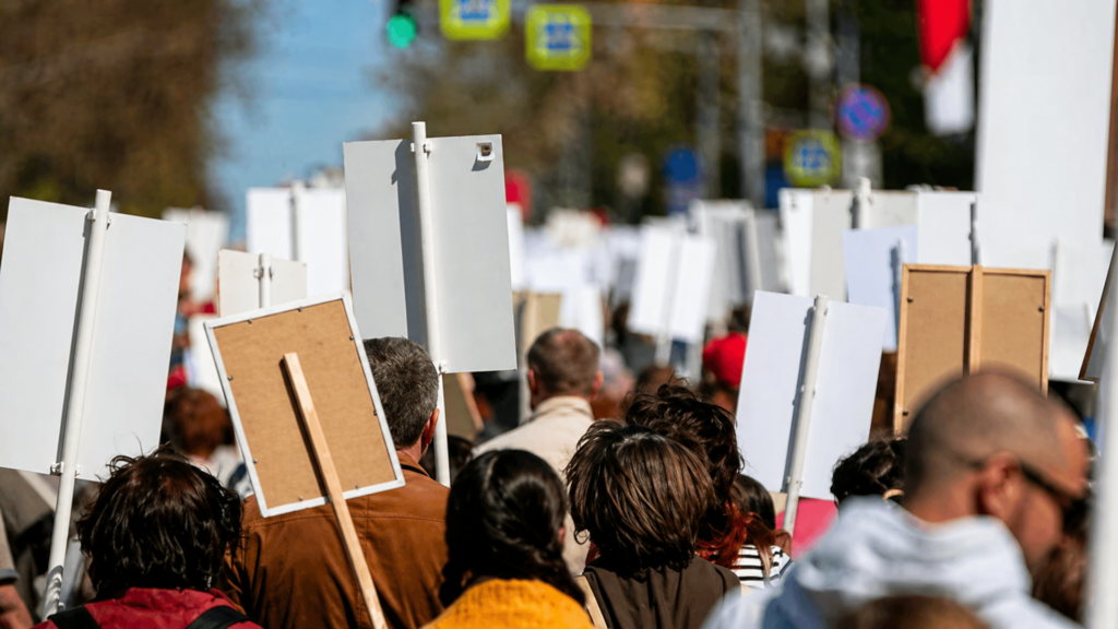 Crowd marches down a street holding protest signs during a daytime demonstration.