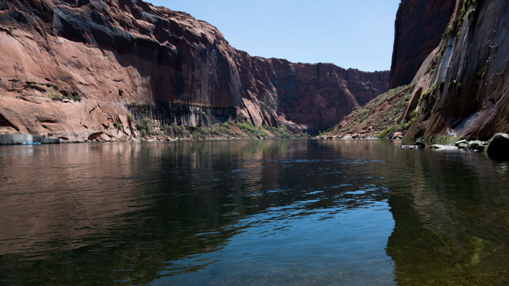 A calm view of the Colorado River reflecting the tall, reddish-brown sandstone cliffs of Glen Canyon under a clear blue sky.