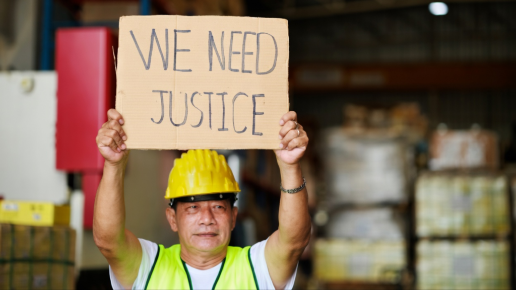 A medium shot of a man in a bright yellow safety vest and hardhat holding up a handwritten cardboard sign.
