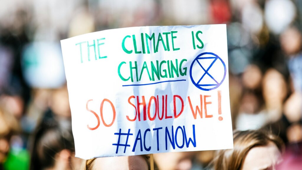 Protester holding sign reading "The climate is changing so should we #ActNow" at demonstration.