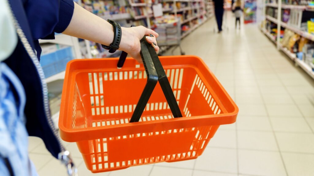 Person carrying an empty shopping basket down a store aisle.