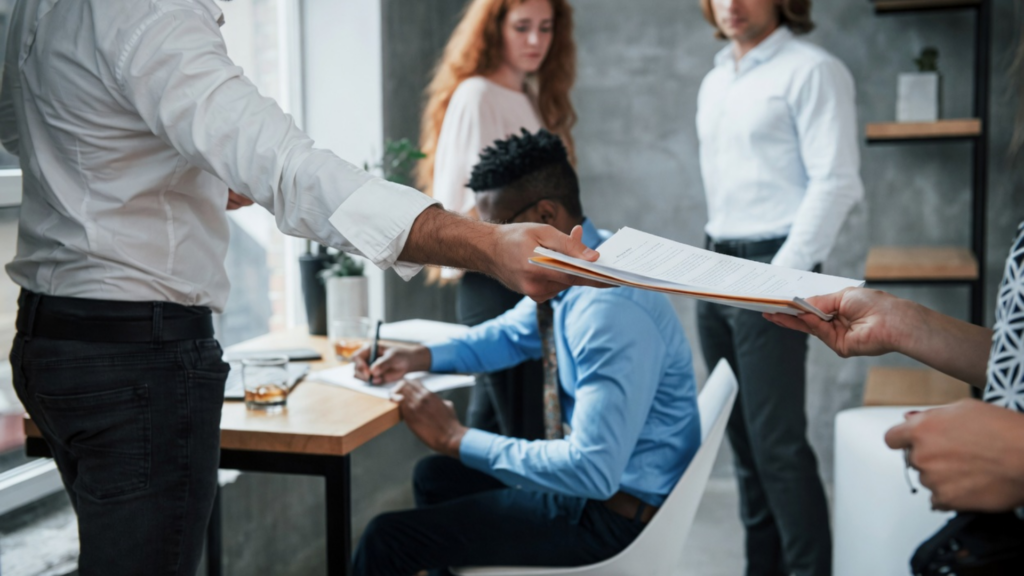 A medium shot of a business setting where one person is handing a white document or legal file to another; several colleagues are blurred in the background, suggesting a formal or legal exchange.