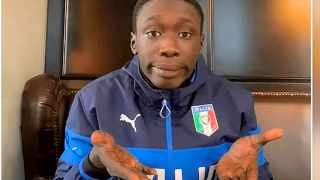 A classic screenshot of Khaby Lame from one of his viral videos, wearing a blue-and-navy Italy national team jacket and performing his signature open-palmed "keep it simple" shrug.
