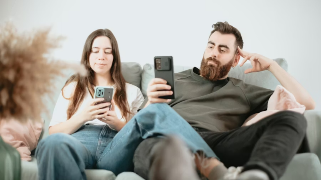 A medium shot of a young man and woman sitting on a light grey sofa, both staring intently at their smartphones; the man has a thick beard and his hand on his head, illustrating the consumption of digital content.