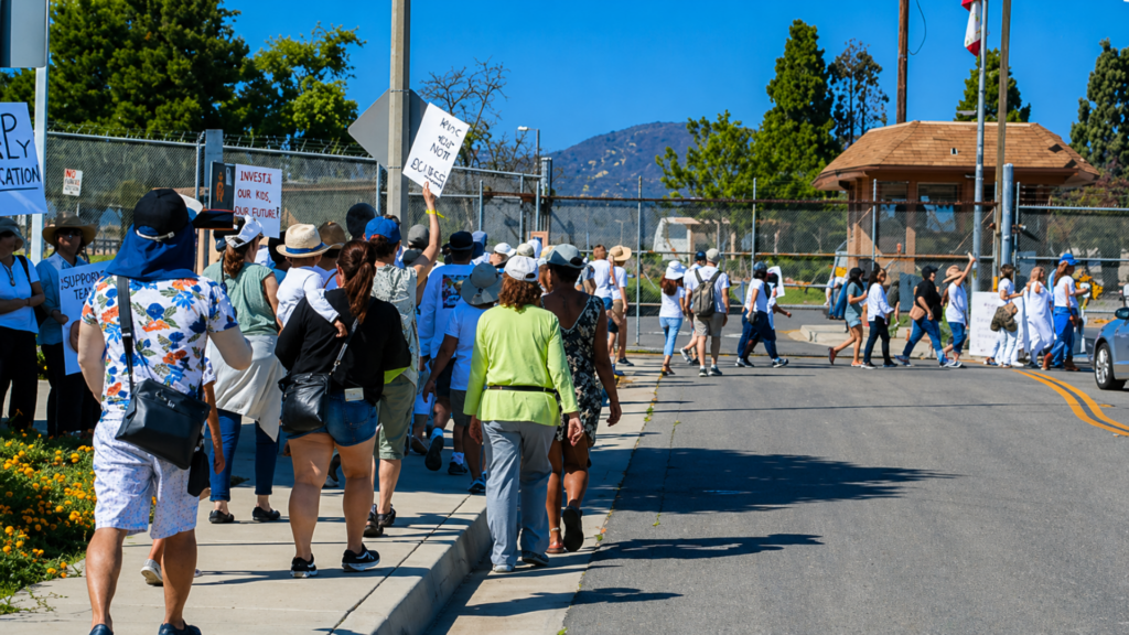 Protesters march along a street holding signs near a fenced facility during a daytime demonstration.