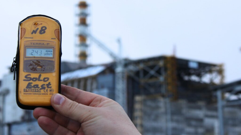 A hand holds a yellow digital dosimeter displaying a radiation reading in the foreground, with the blurred structure of the Chernobyl nuclear power plant and a crane in the background.