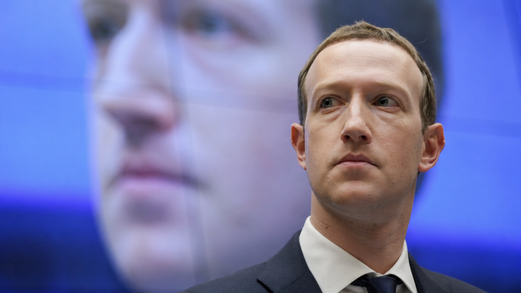 Close-up portrait of Mark Zuckerberg in a dark suit, looking off-camera, with a large digital screen behind him displaying a magnified version of his face.