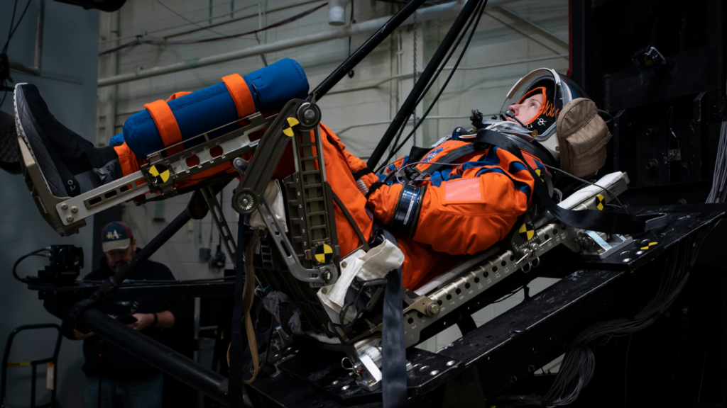 An astronaut in a bright orange flight suit lies reclined in a specialized testing chair while wearing a helmet and mission gear.
