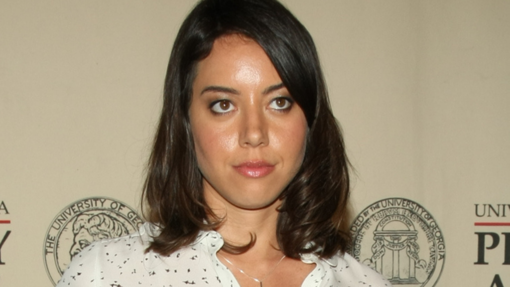 Aubrey Plaza stands against a tan backdrop wearing a white patterned blouse.