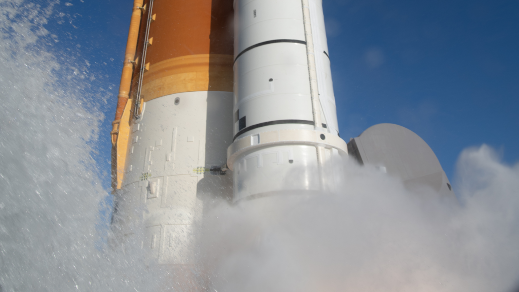 The massive orange and white Artemis II rocket engines ignite, creating a thick plume of white smoke and bright orange flames during liftoff.