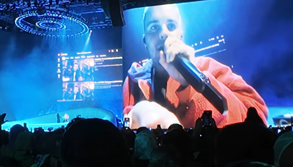 Justin Bieber singing into a microphone as his old YouTube videos play on a large screen during his Coachella 2026 set.