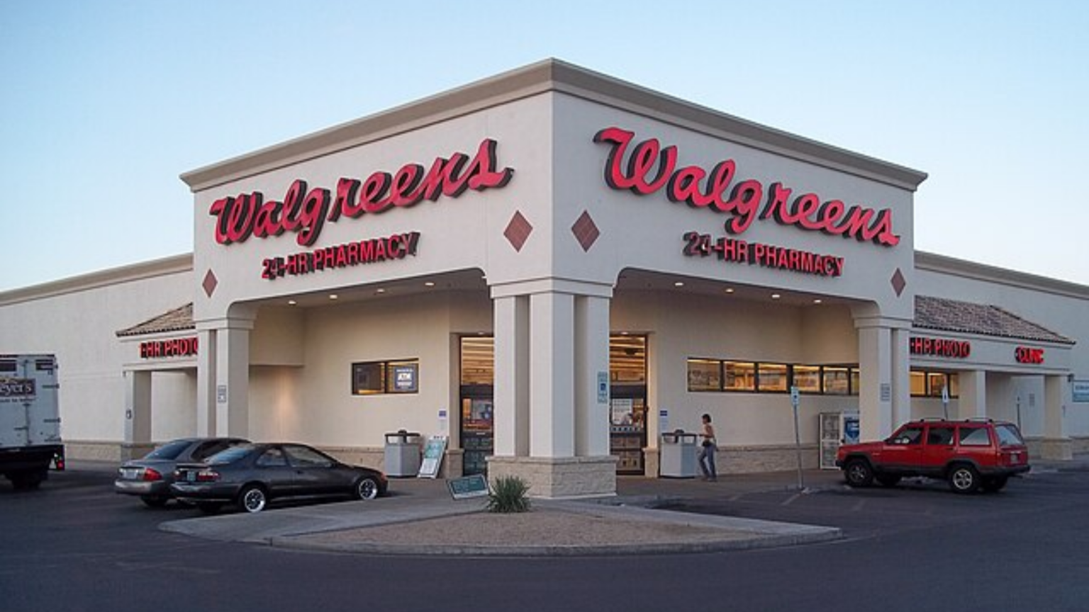 A front-angle view of a white Walgreens pharmacy building with its signature red cursive logo against a pale sky.