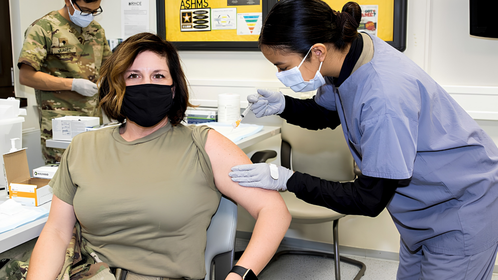 A healthcare worker in blue scrubs and a face mask administers a vaccine to a woman wearing a military undershirt and a black face mask.