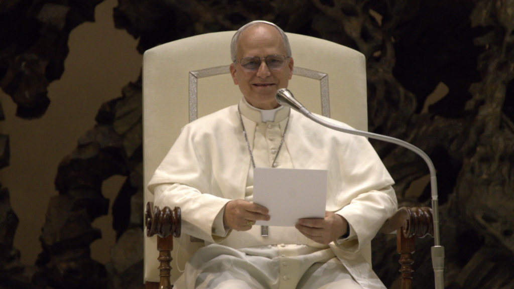 A man in white papal attire and glasses sits in a large cream-colored chair holding a white paper.