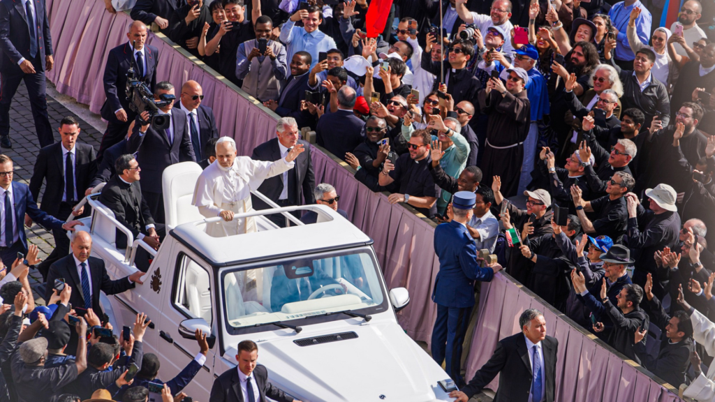 A newly inaugurated Pope waves to a cheering crowd from the back of a white popemobile as it drives through a sunlit city street.