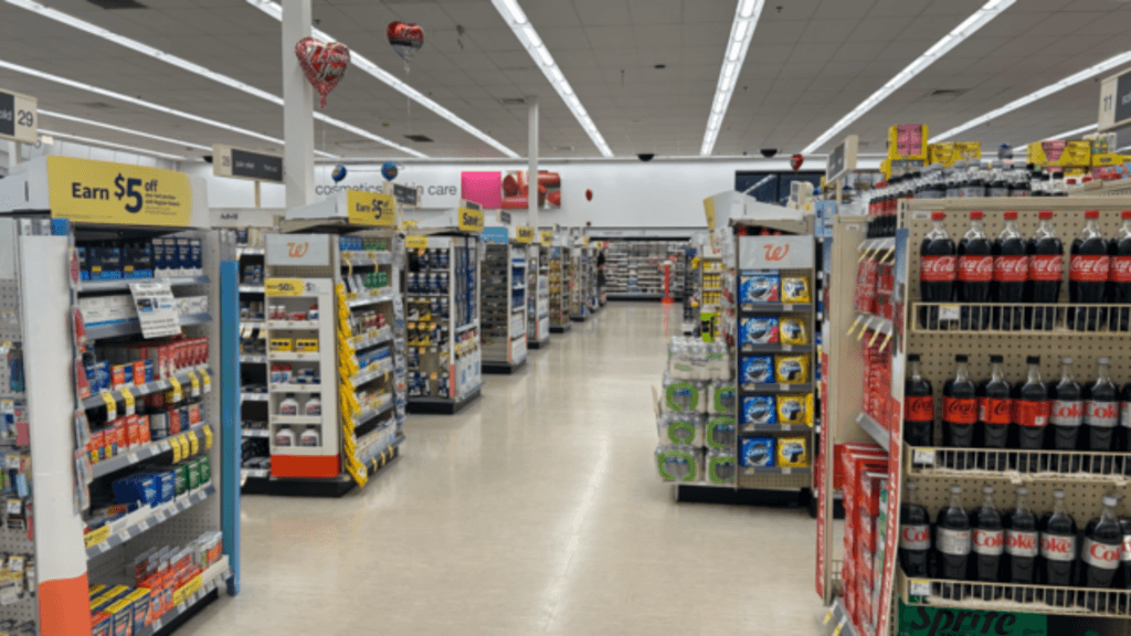 An interior view of a Walgreens store showing long, brightly lit aisles stocked with beverages, snacks, and health products.