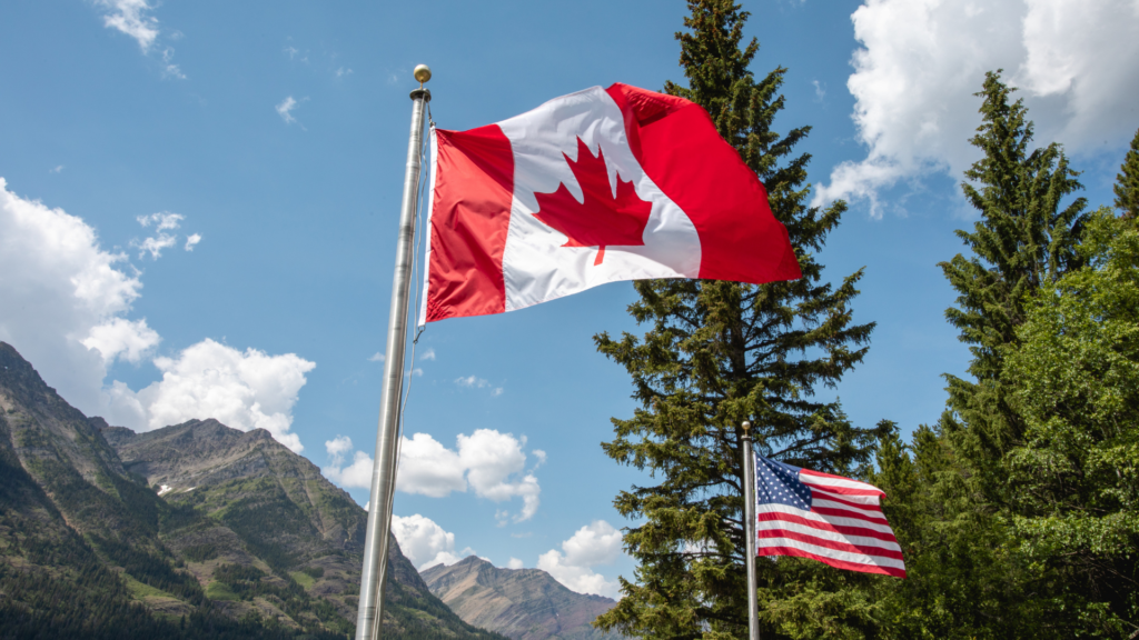 The Canadian and American flags fly high on separate poles against a backdrop of towering evergreen trees and rugged, sunlit mountains.