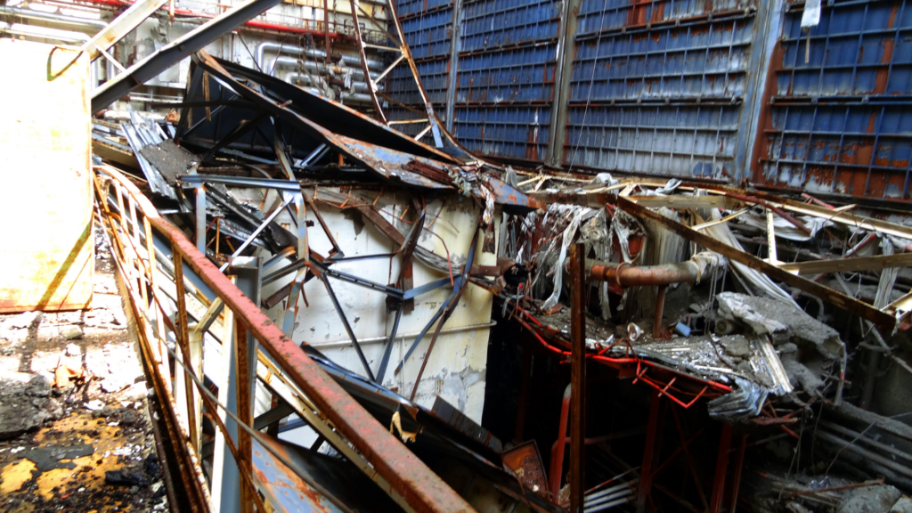 Twisted metal beams, debris, and rusted industrial structures clutter the interior of a heavily damaged and abandoned turbine hall.