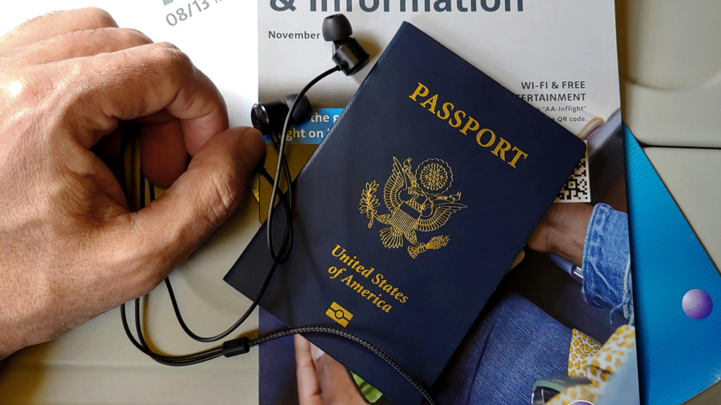 A blue United States passport rests on an in-flight magazine next to a pair of black wired earbuds and a person's hand.