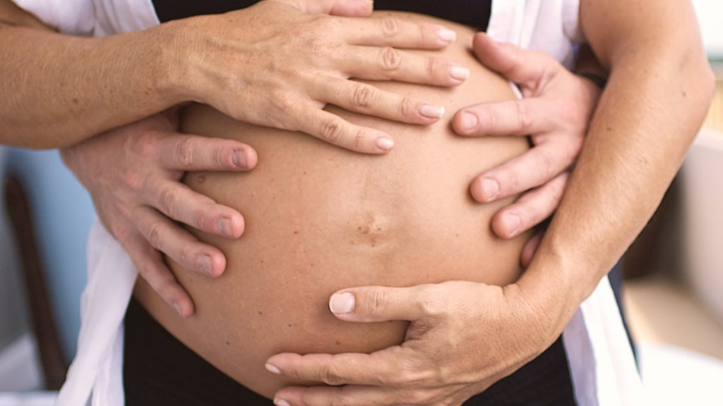 A close-up, top-down view of several hands resting gently on a pregnant person's bare stomach.