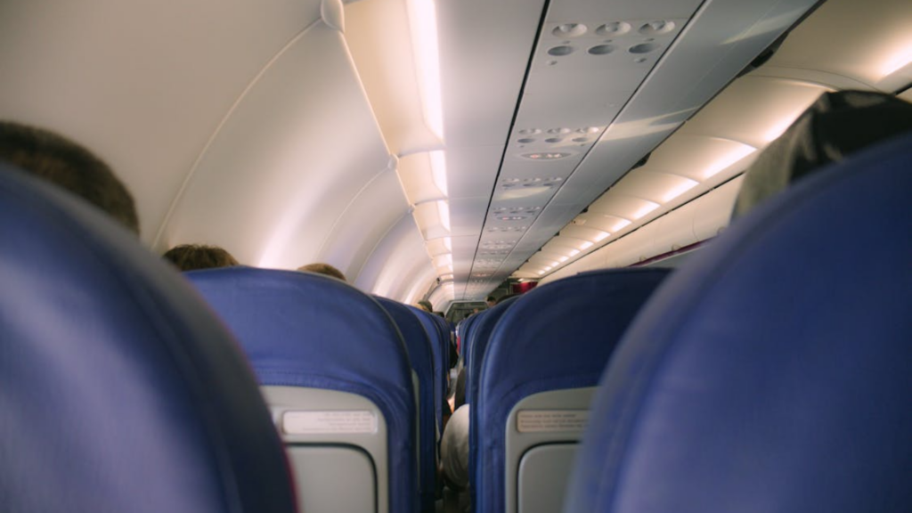 A view down the aisle of a commercial airplane cabin, showing the tops of blue seats and the overhead luggage bins.