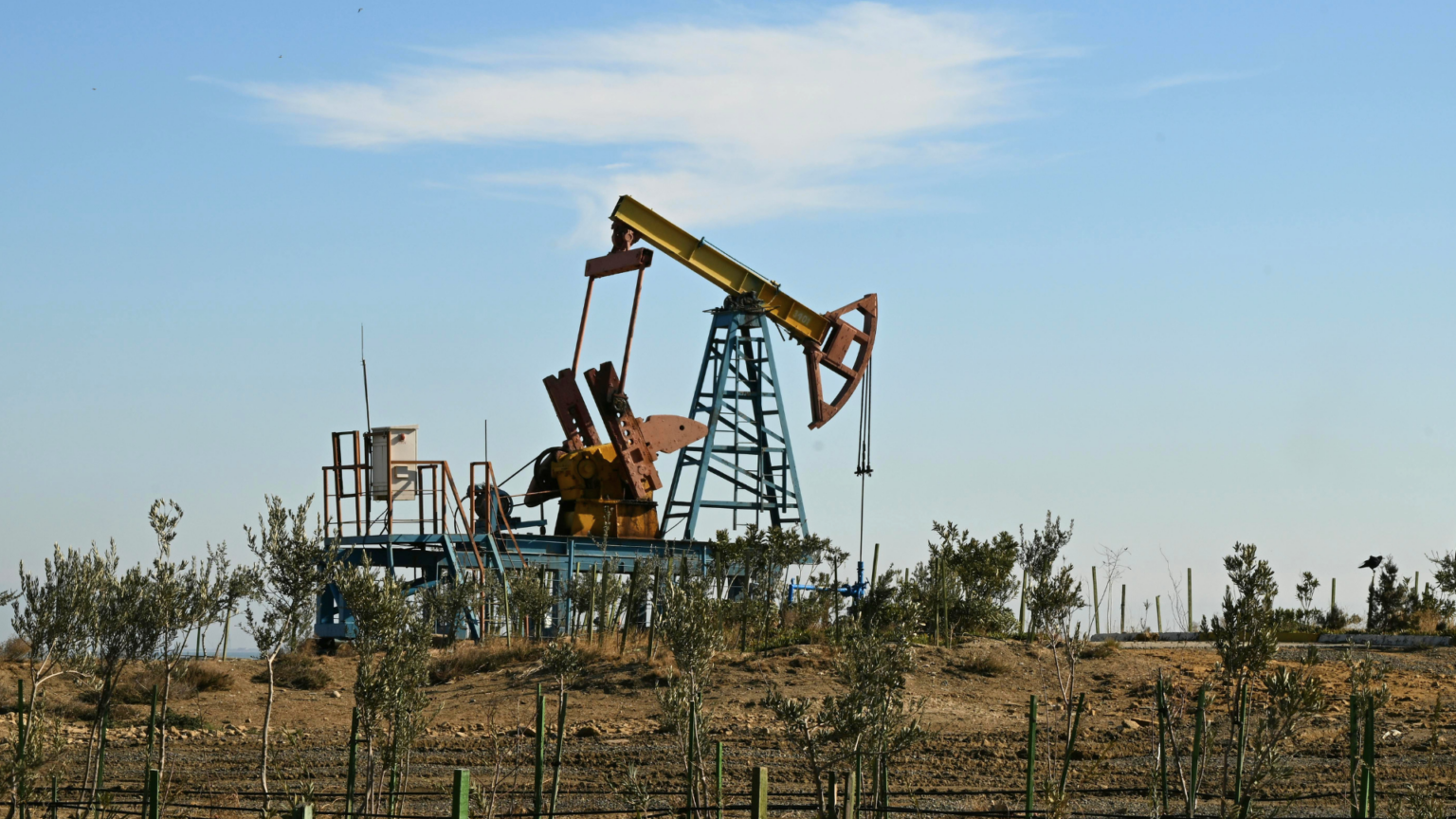 An industrial oil pump jack stands on a flat, arid landscape under a clear blue sky with a few wispy white clouds.