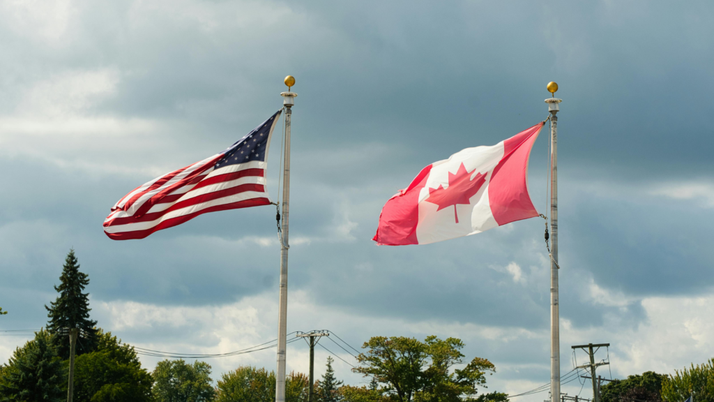 The American and Canadian flags wave side-by-side on tall flagpoles under a vast, cloudy sky overlooking a line of distant trees.