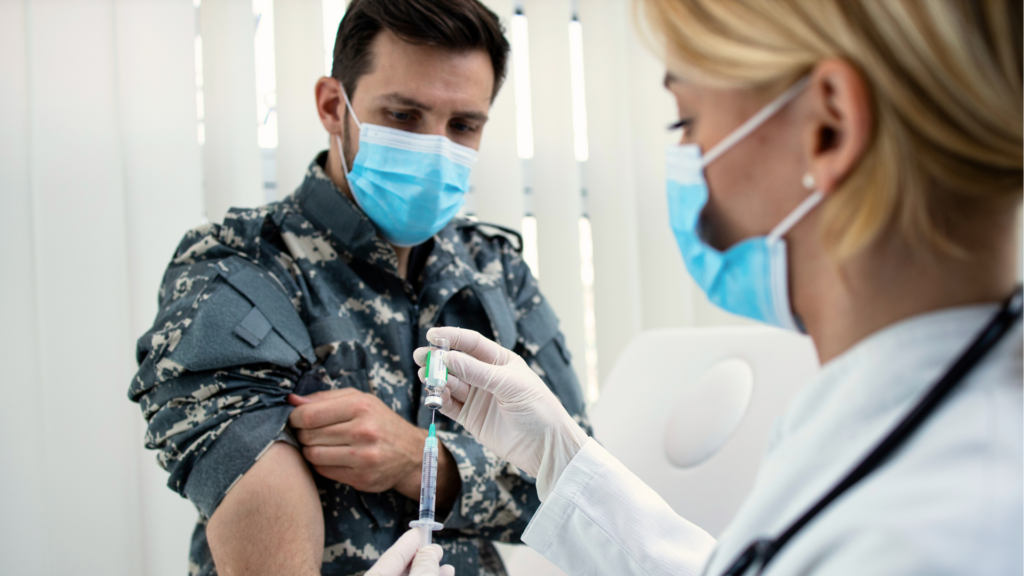 A healthcare provider in a mask and white coat prepares a syringe to administer a vaccine to a service member in a camouflage uniform.