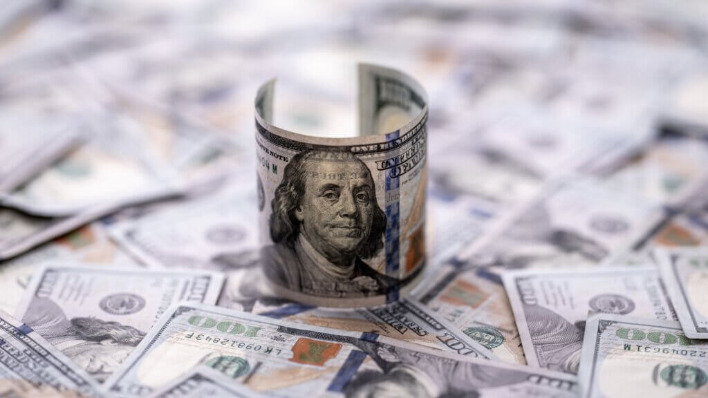 A rolled-up 100-dollar bill stands upright among a scattered pile of similar bills.