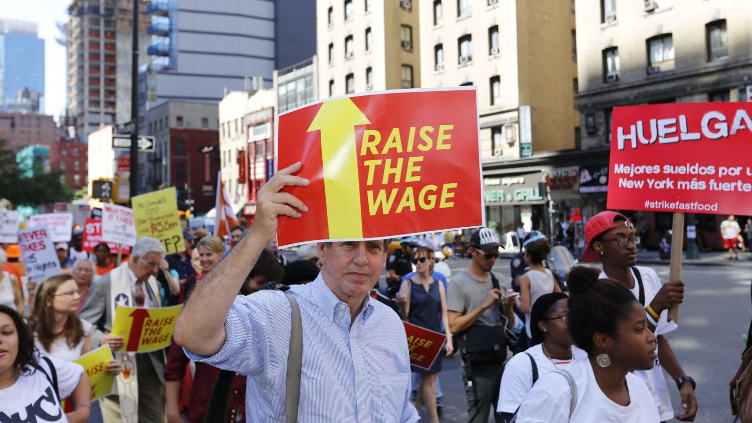 Protesters marching through a New York City street holding 'RAISE THE WAGE' signs.