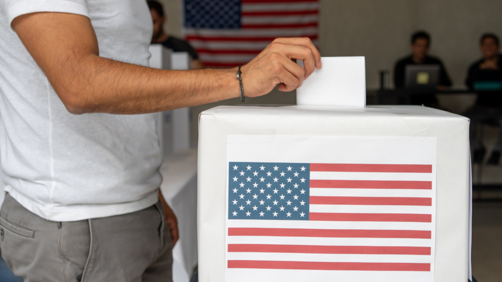 A person in a white t-shirt placing a paper ballot into a white ballot box featuring the American flag.