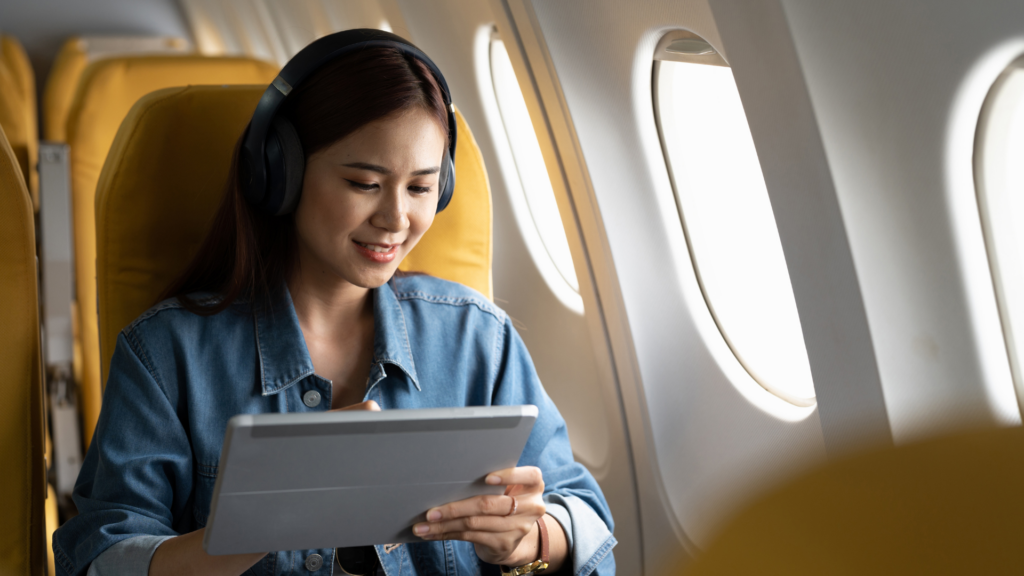 A woman wearing black headphones sits in an airplane seat, smiling while using a silver tablet.