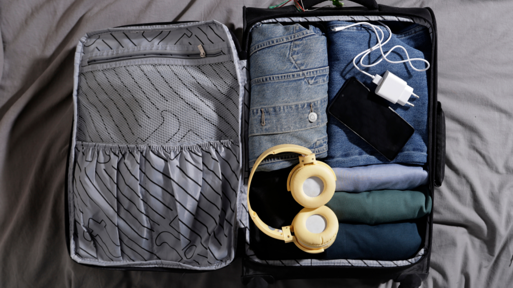 An open black suitcase on a gray bedspread, neatly packed with folded denim, t-shirts, a phone, a charging block, and yellow headphones.